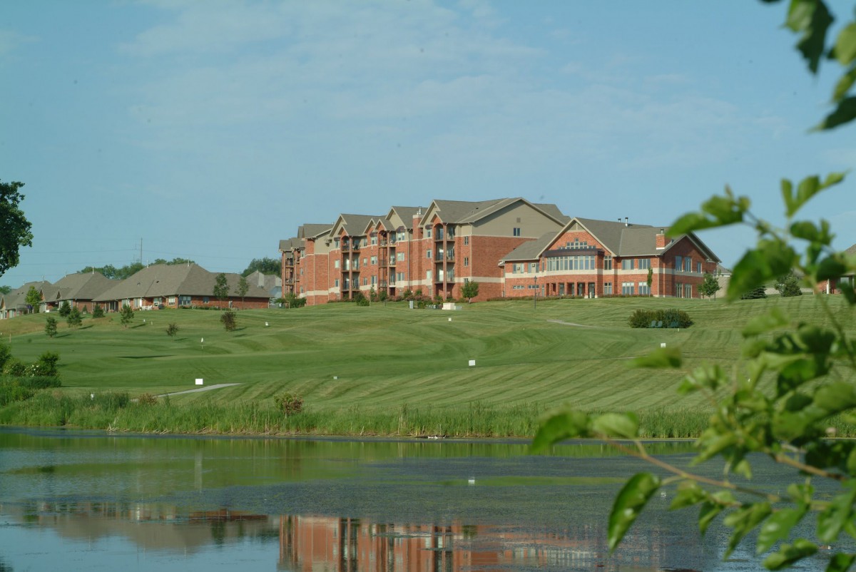 A large building sits by a calm lake, surrounded by green grass and under a blue sky.