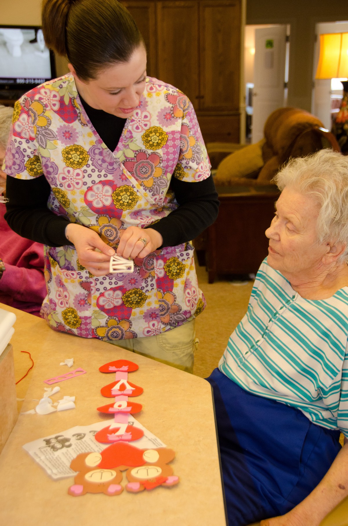 A caregiver assists an elderly woman with a craft activity in a nursing home.