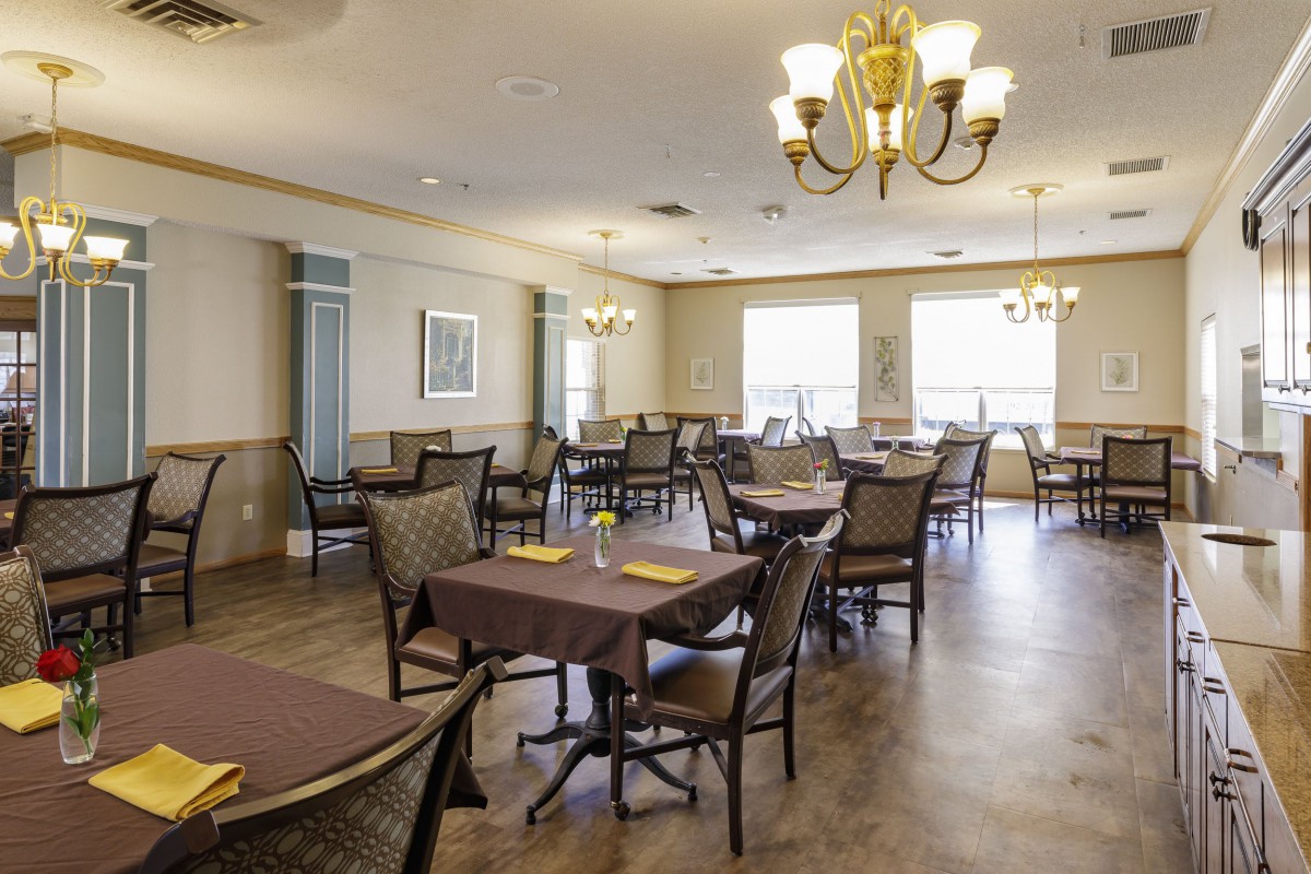 An empty dining room with neatly arranged tables and chairs, illuminated by chandeliers and natural light from large windows.