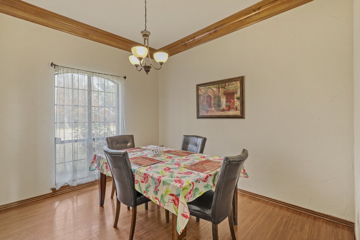 A well-lit dining area with a table covered in a floral tablecloth, surrounded by four chairs. A chandelier hangs above and a painting decorates the wall.