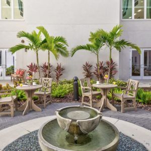 A serene courtyard featuring a central fountain, surrounded by tables and chairs, with palm trees and plants enhancing the tranquil atmosphere.