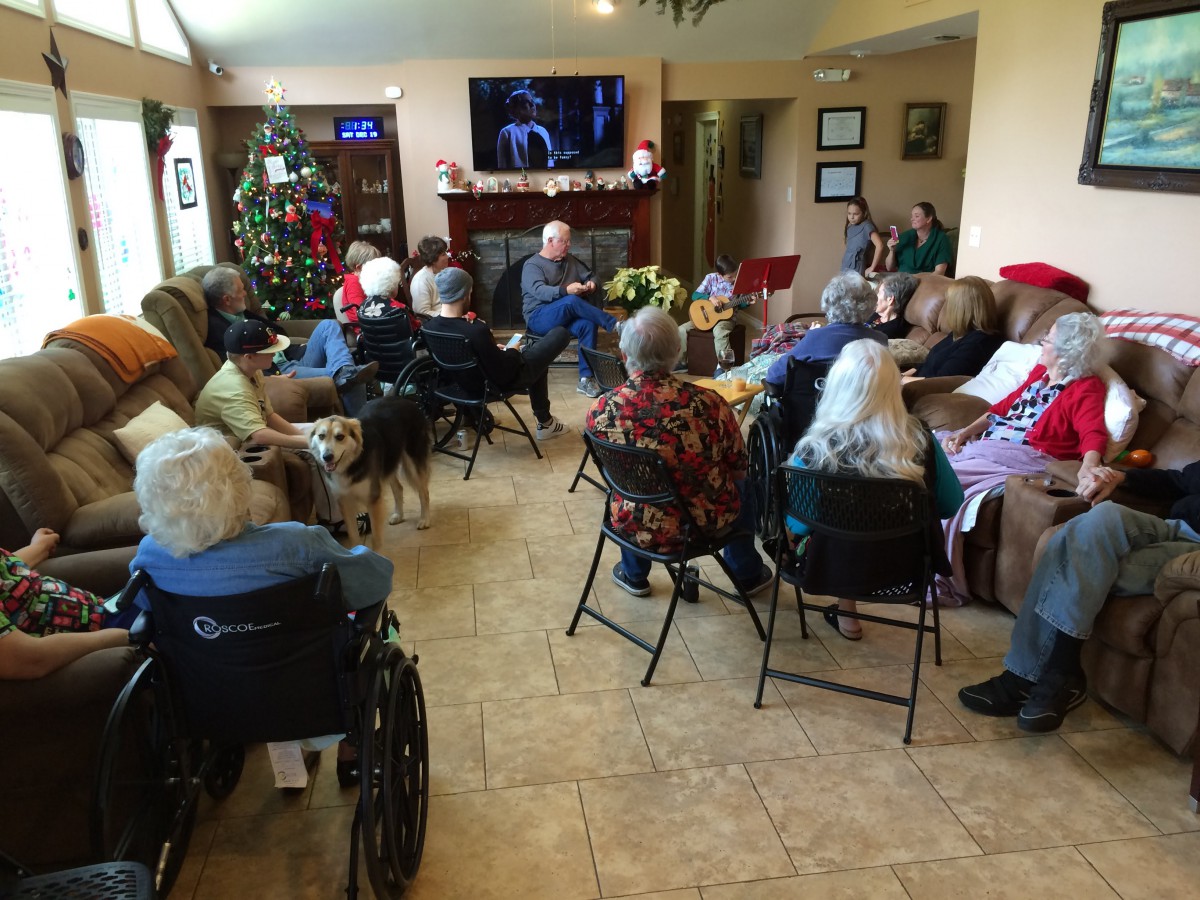 A group of elderly individuals gathered in a common room, watching television with festive holiday decorations around.