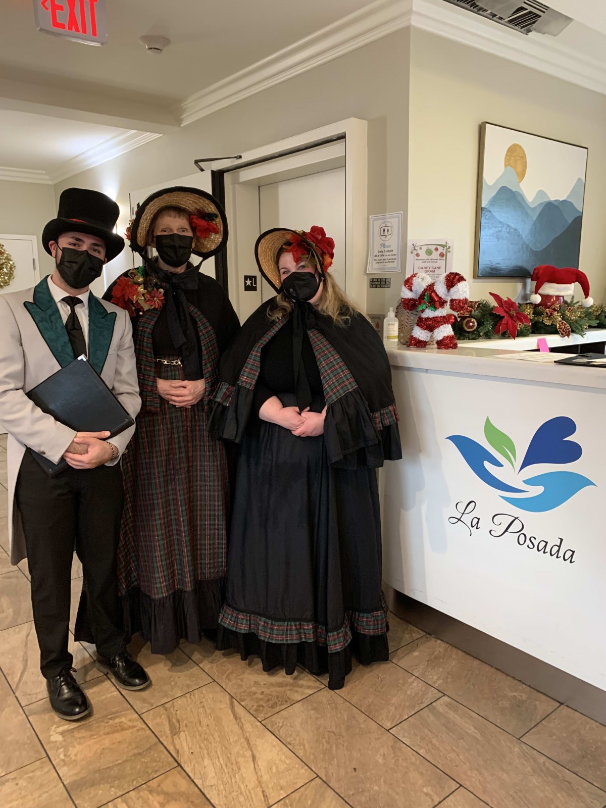 Three people in period costumes and masks stand by a reception desk.