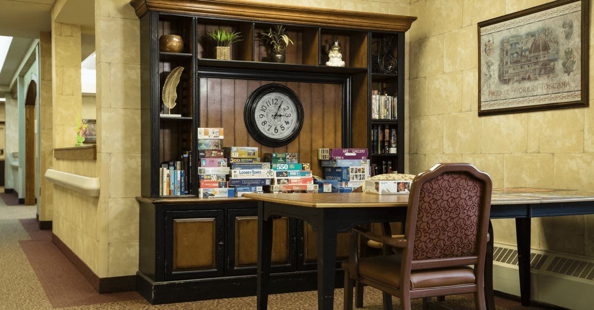 A cozy interior scene featuring a wooden bookshelf filled with books and board games, a wall clock, and a wooden table with chairs.