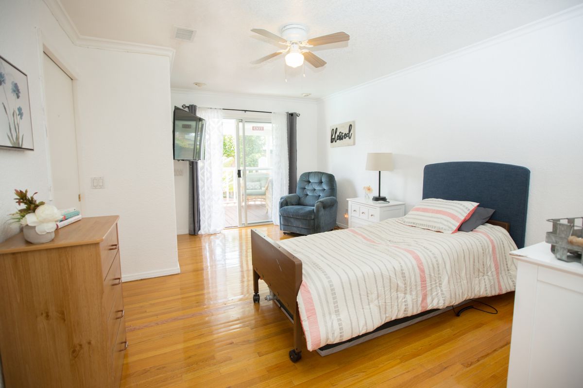 A bright bedroom with a bed, armchair, and a ceiling fan, featuring hardwood floors and sliding glass doors.