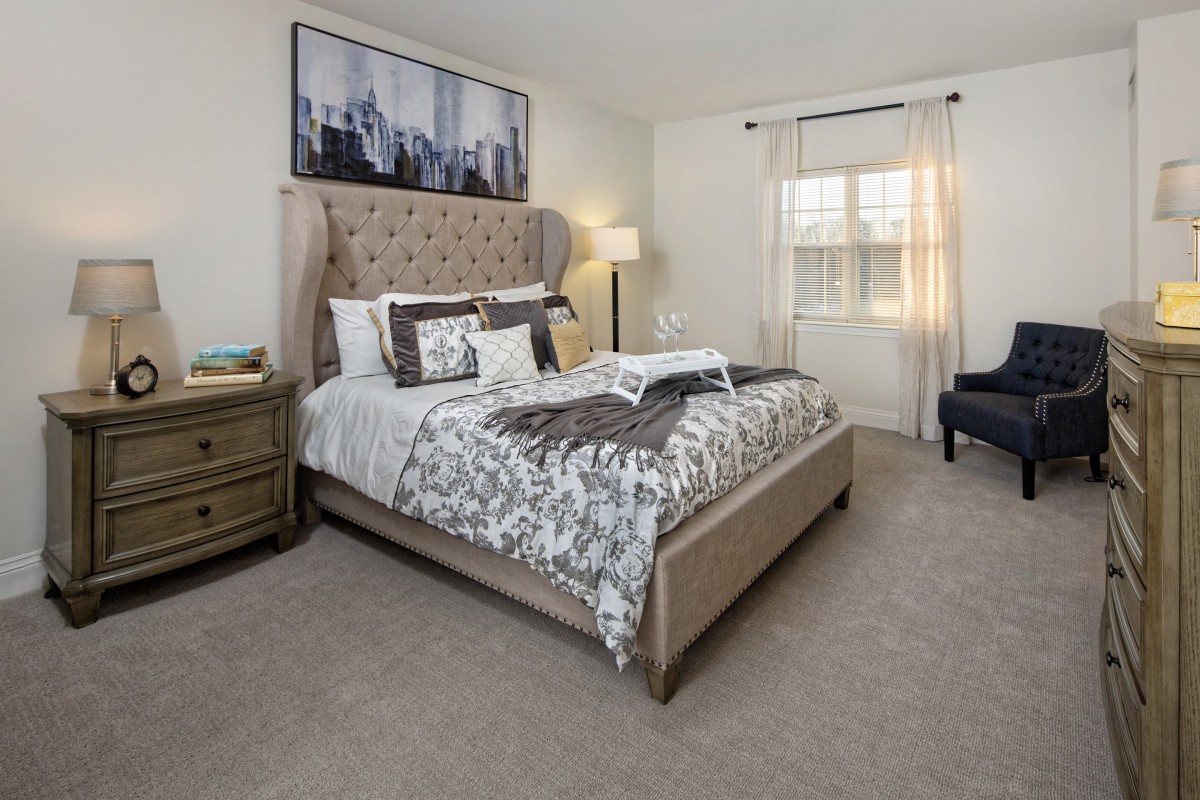 A cozy and elegantly decorated bedroom featuring a plush bed, bedside table, floor lamp, and a window with light curtains.