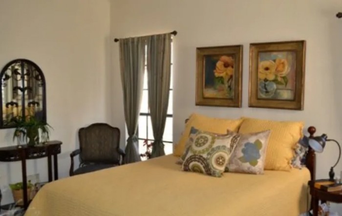 A cozy bedroom with a yellow bedspread and decorative pillows, featuring artwork on the wall and curtains by the window.