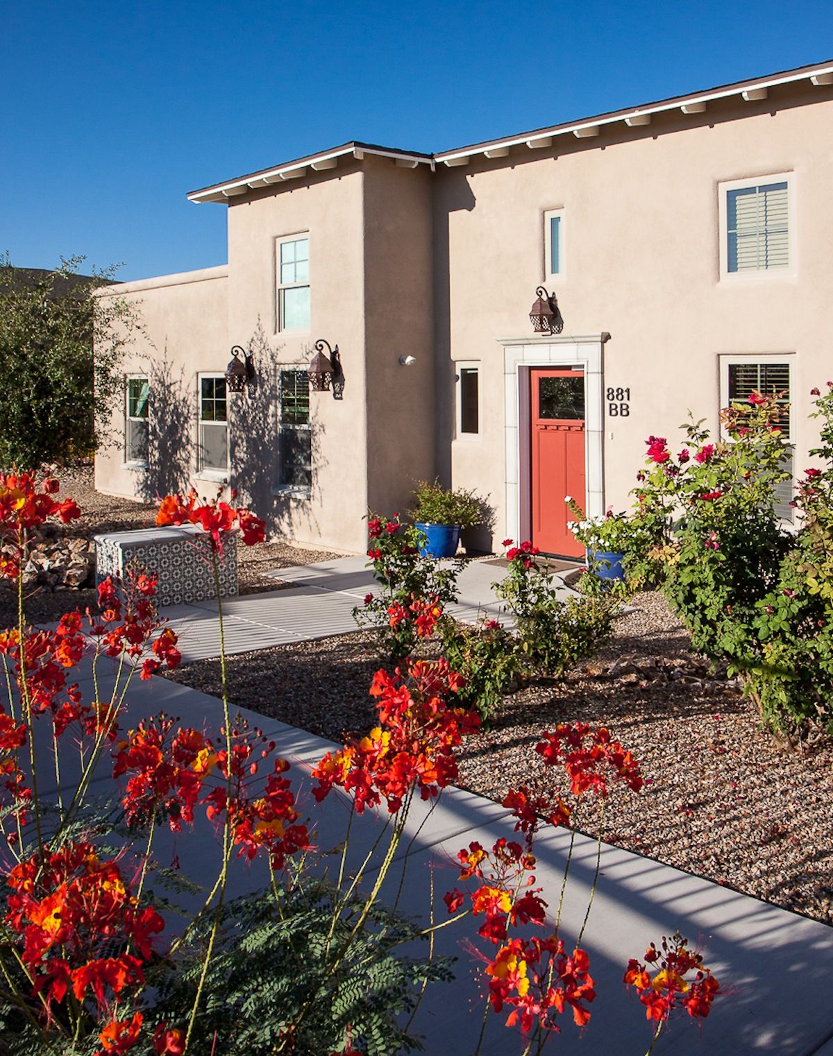 A stucco house with a vibrant garden featuring red and orange flowers in a desert landscape.