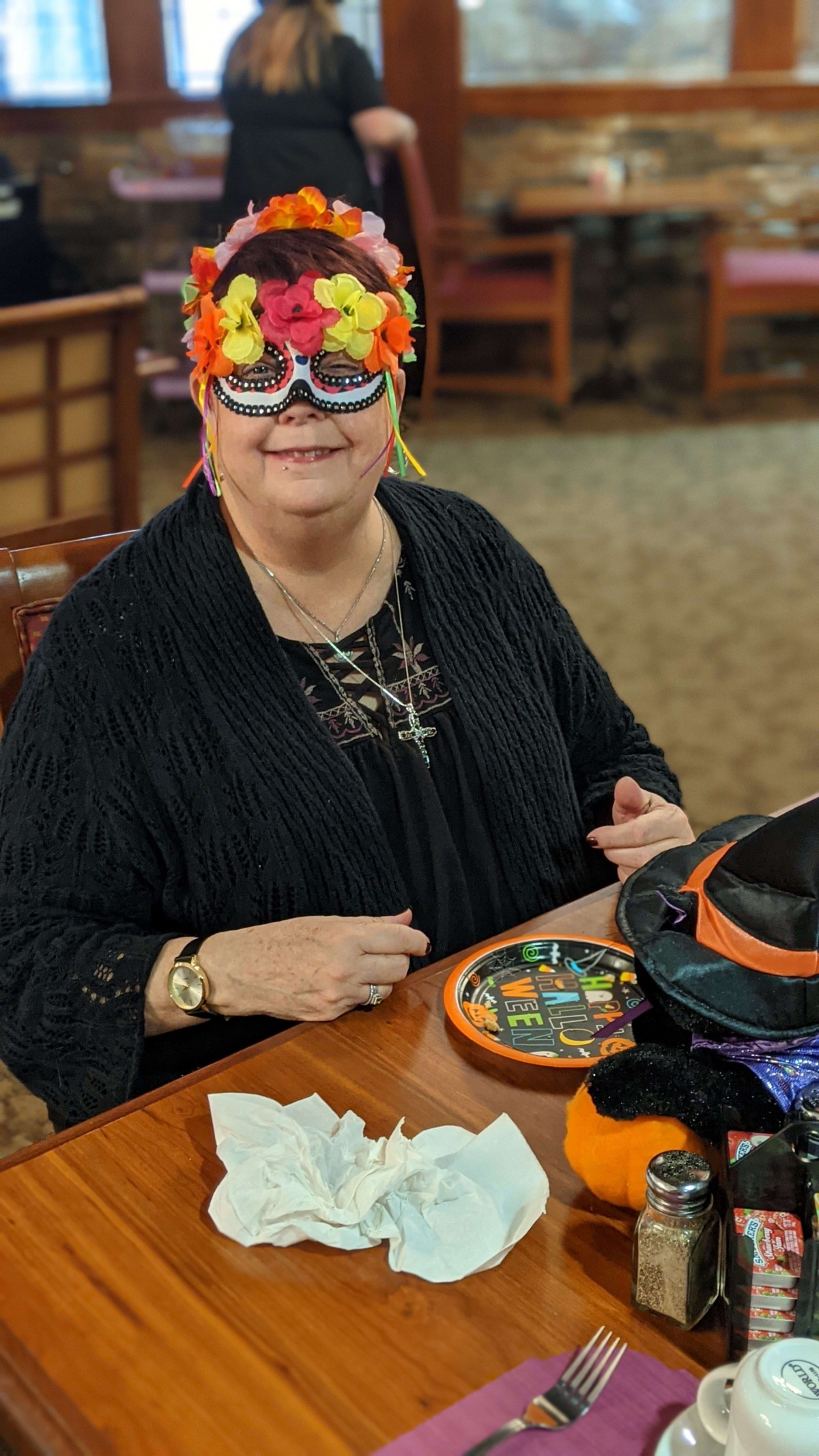 A person wearing a colorful mask is smiling while sitting at a table with festive decorations.