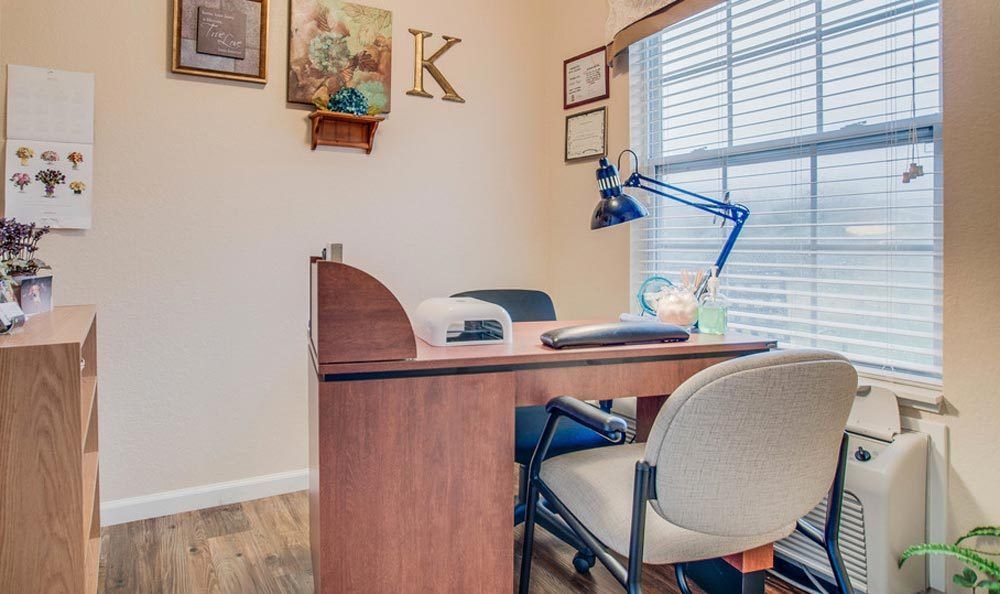 A neatly arranged manicure station inside a nail salon with a desk, lamp, and chairs by a window.