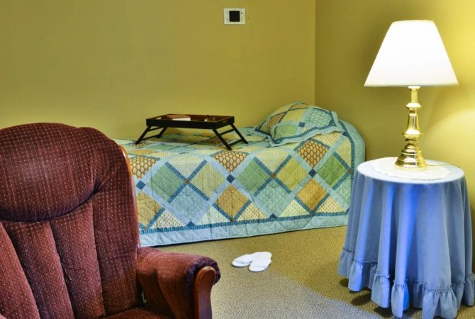 A cozy bedroom with a patterned quilt on the bed, an armchair, and a nightstand with a lamp.
