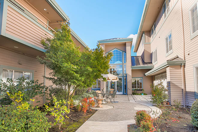 A view of a modern building's courtyard featuring trees, plants, and outdoor furniture under a clear blue sky.
