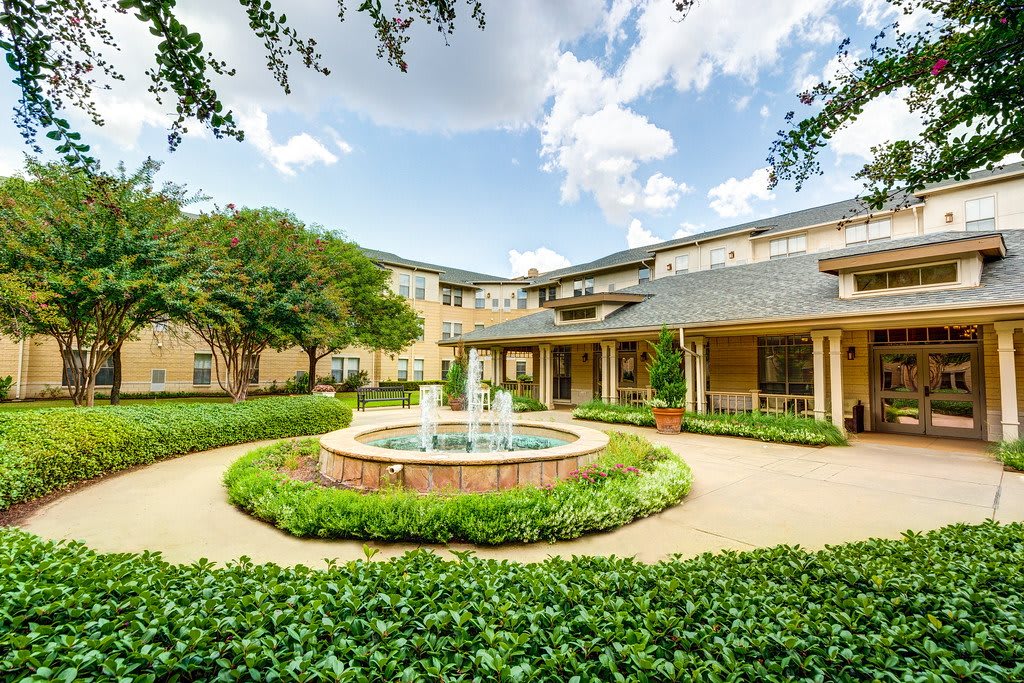 A landscaped garden with a central fountain surrounded by greenery and a two-story building under a blue sky.