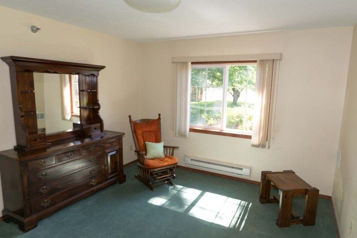 A cozy room featuring a wooden rocking chair, an ornate dresser, and a large window letting in natural light.