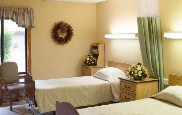 A simple hospital room featuring two twin beds, adorned with floral arrangements and a wall-mounted wreath.