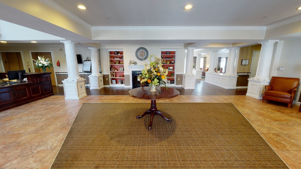 A spacious lobby with a central wooden table displaying a flower arrangement, surrounded by bookshelves and elegant decor.