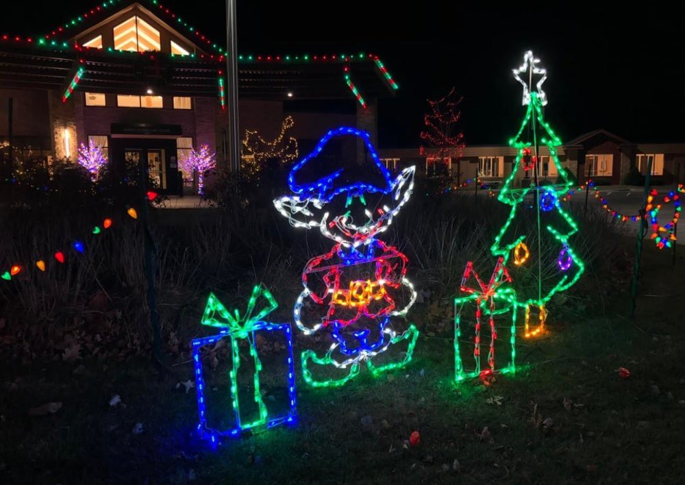 A festive holiday display featuring an illuminated elf holding a gift, surrounded by colorful presents and a Christmas tree.
