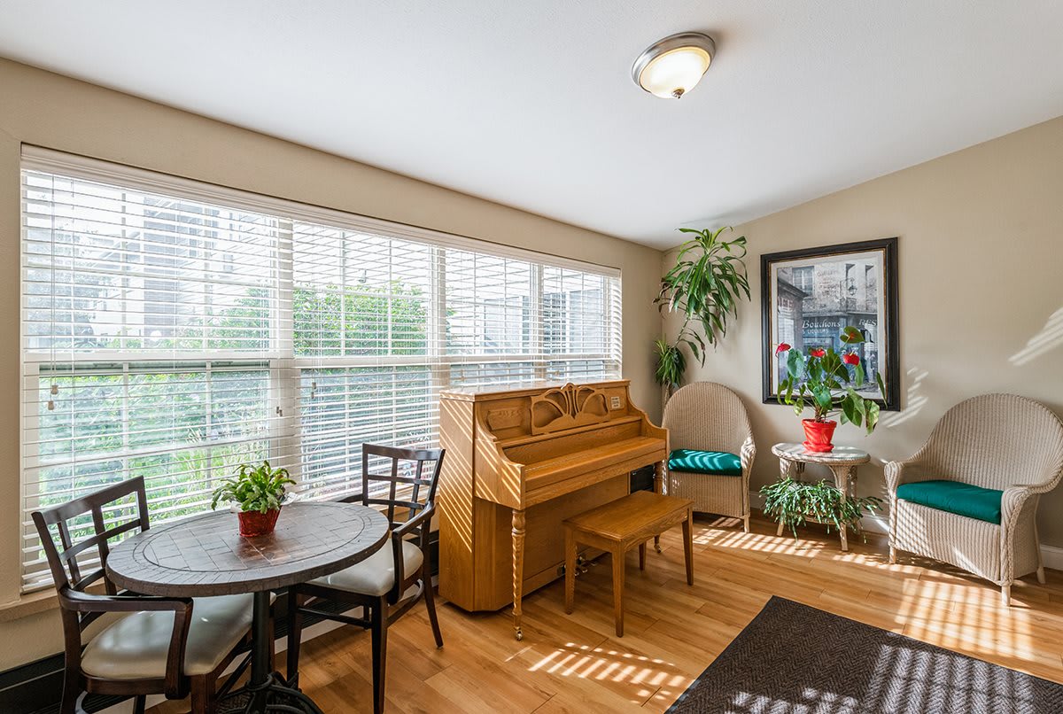 A cozy living room filled with natural light, featuring a piano and a small dining set surrounded by lush plants.