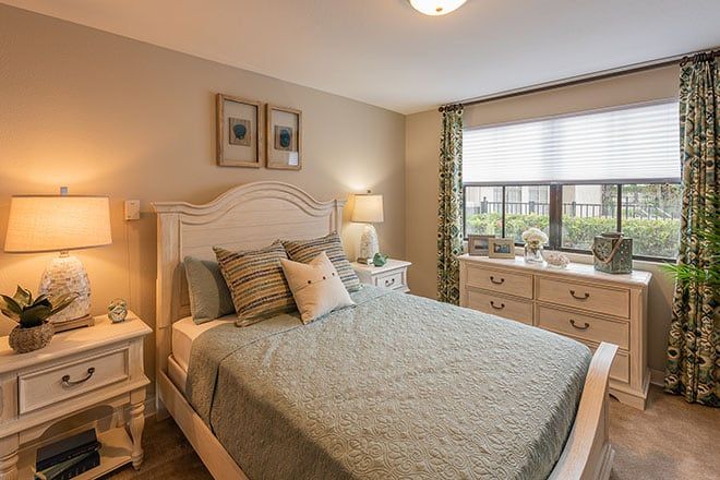 A cozy bedroom interior featuring a neatly made bed, bedside tables with lamps, and a window with patterned curtains.