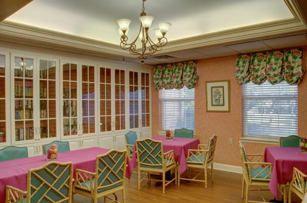 A cozy dining area with colorful tablecloths and a chandelier, featuring large windows and decorative curtains.