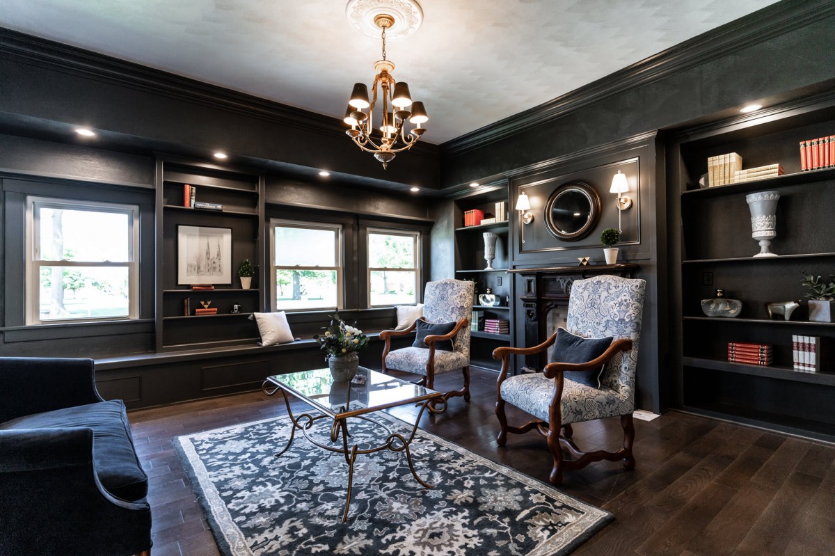 A traditional living room with dark walls, featuring elegant armchairs, a rug, and a chandelier.