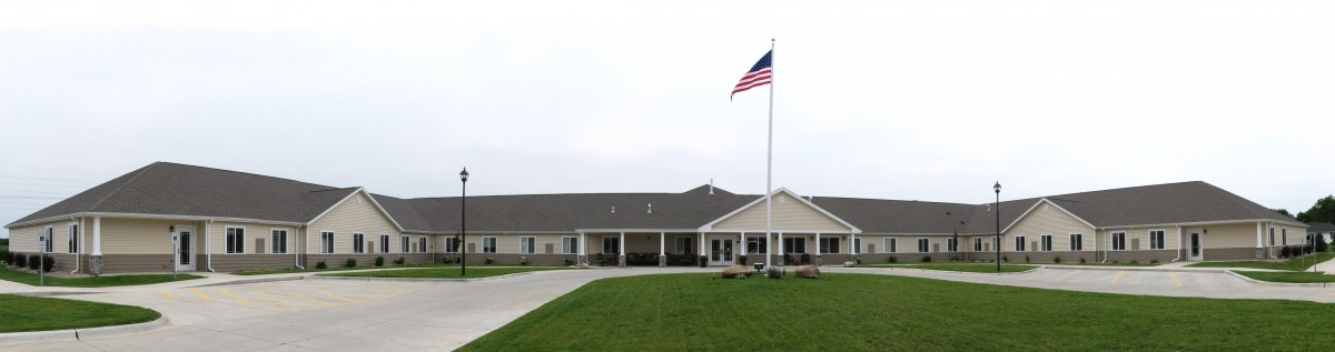 A building with an American flag on a tall pole, surrounded by a green lawn and driveway under an overcast sky.