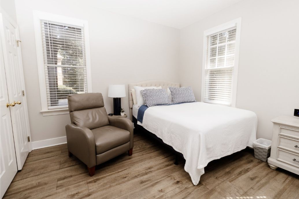 A minimalist bedroom with a bed, armchair, and lamp, featuring large windows and hardwood flooring.