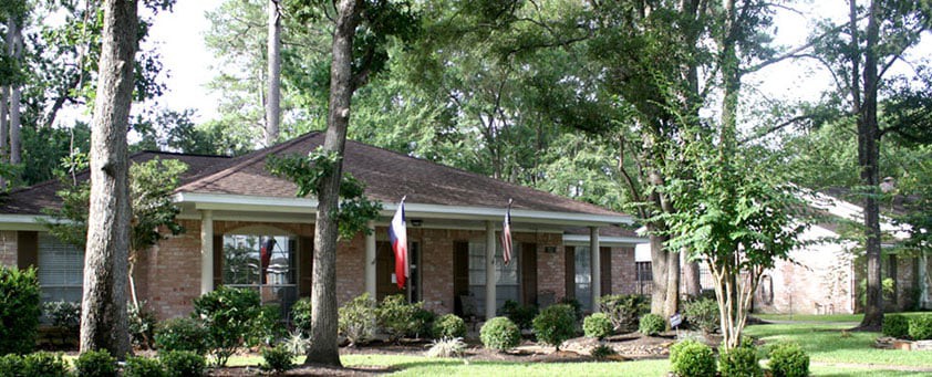 A suburban brick house surrounded by trees and a well-maintained garden, with flags displayed at the front.