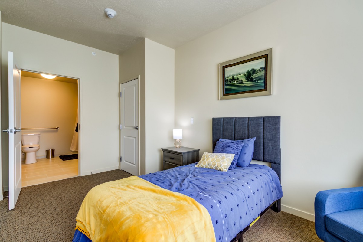 A cozy bedroom with a single bed featuring blue bedding and a yellow blanket, complemented by a nightstand and lamp, all set on a carpeted floor.