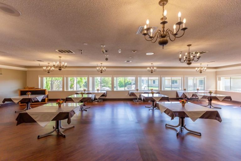 A spacious dining room with chandeliers, several tables, large windows, and a wooden floor.
