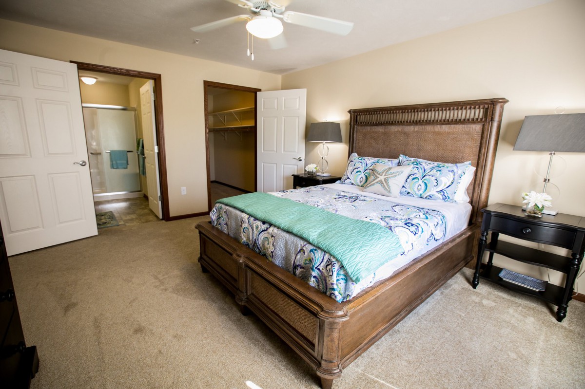 A cozy bedroom with a wooden bedframe, colorful bedding, a nightstand with a lamp, and an open closet door, featuring a ceiling fan for comfort.