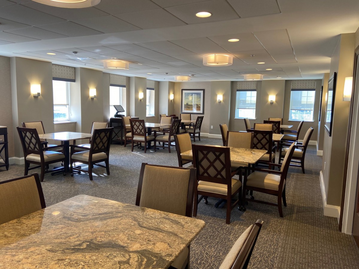 A spacious dining area with multiple tables and chairs, featuring large windows and warm lighting.