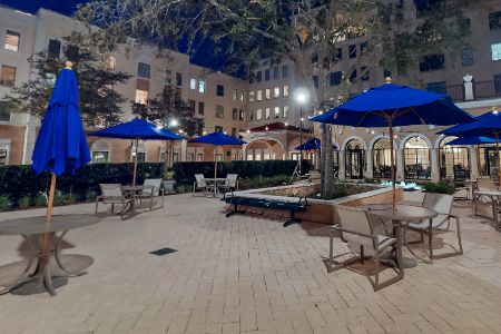 A nighttime view of a well-lit outdoor patio area with tables and chairs under blue umbrellas, surrounded by buildings.