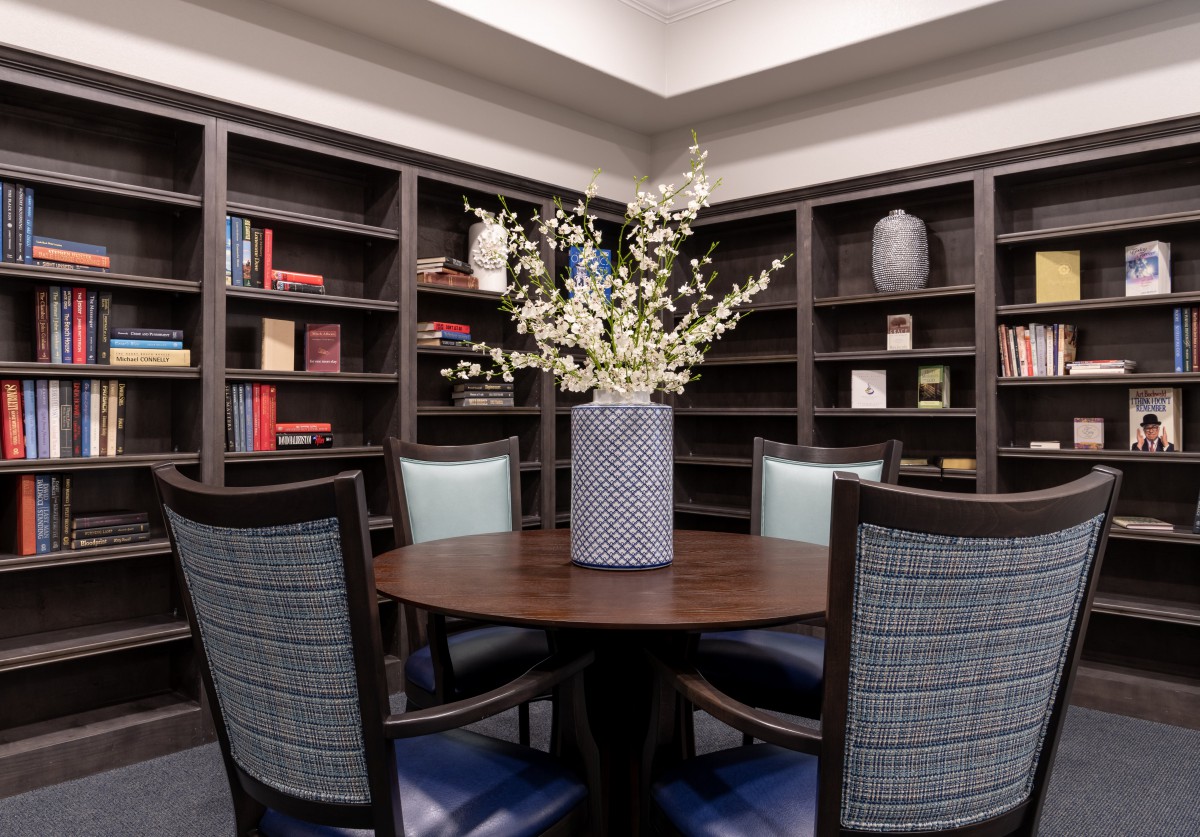 A cozy library corner with a round table and chairs, surrounded by bookshelves, featuring a decorative vase with flowers.