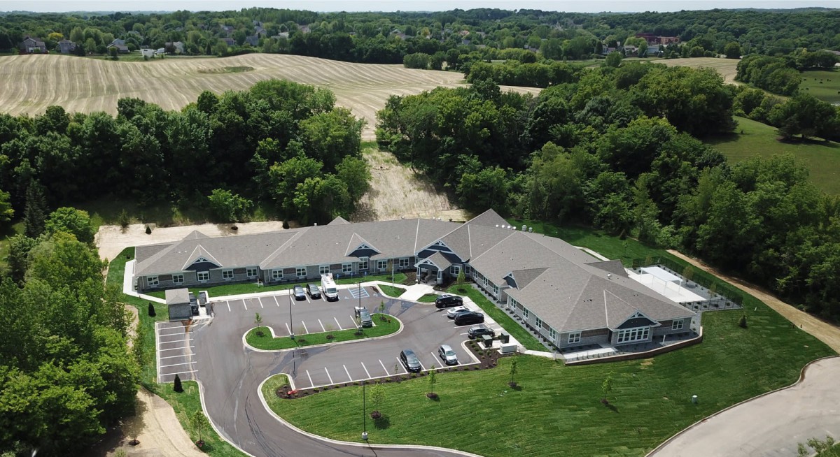 An aerial view of a building with an adjacent parking lot set in a rural landscape surrounded by trees.