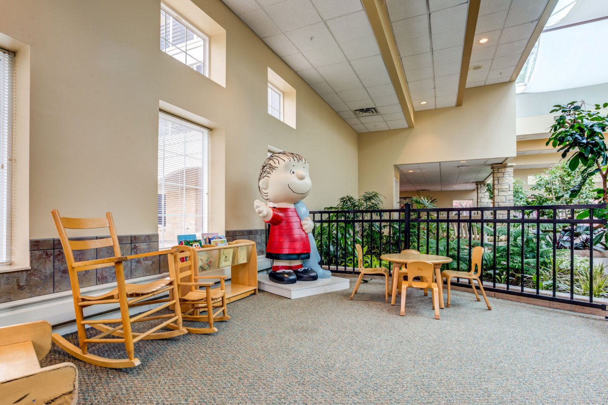 A children's play area featuring a large character statue, small tables and chairs, wooden rocking chairs, and windows allowing natural light.