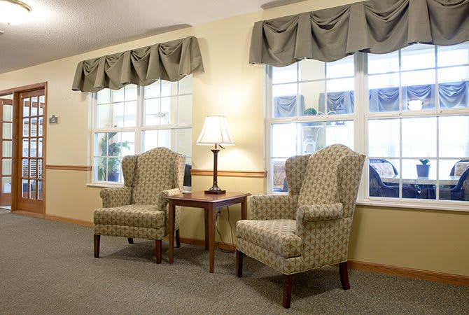 A cozy living room with two upholstered chairs, a side table, and a lamp near large windows.