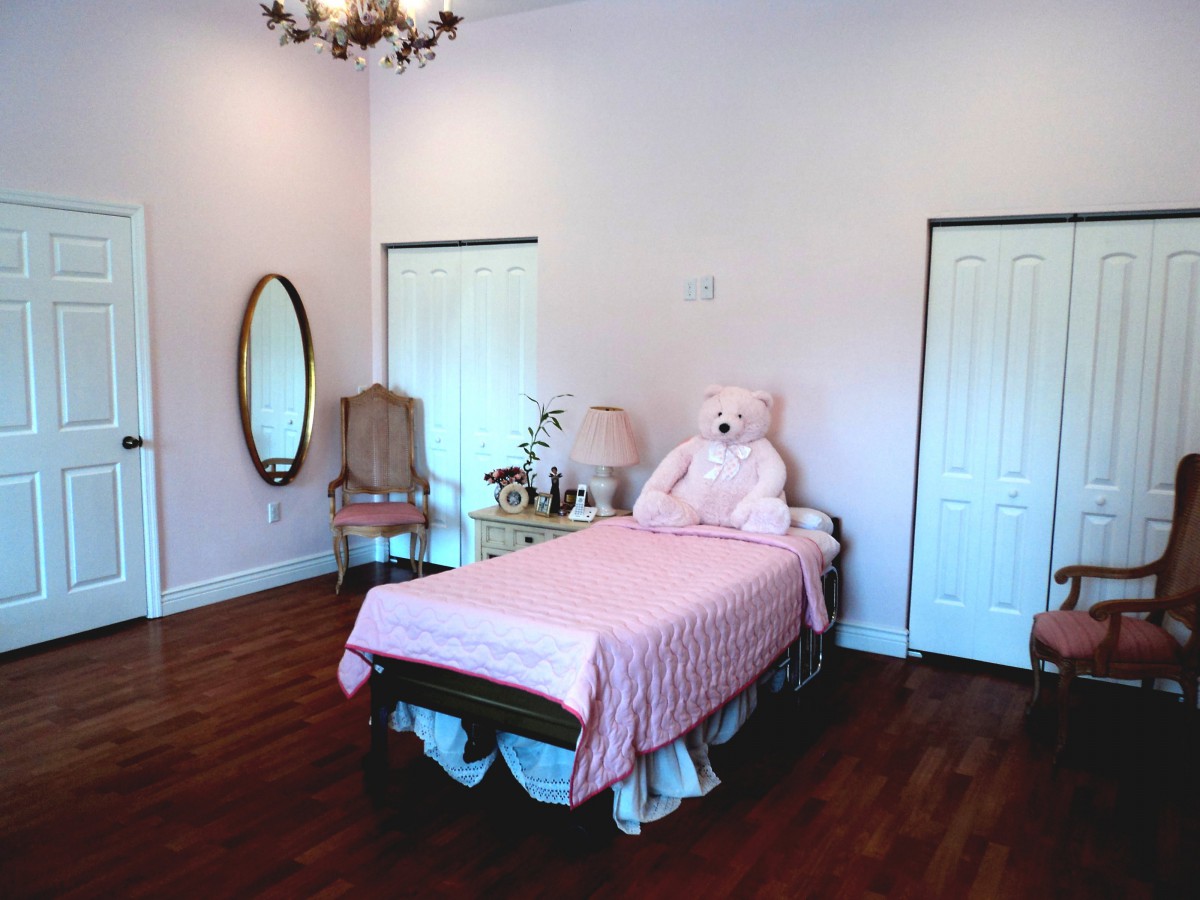 A cozy bedroom with a pink theme, featuring a single bed adorned with a large teddy bear and decorative pillows, complemented by elegant furniture and a chandelier.