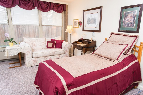 A cozy bedroom with elegant decor, featuring a bed with red and beige bedding, a small wooden desk, and a sofa near the window.
