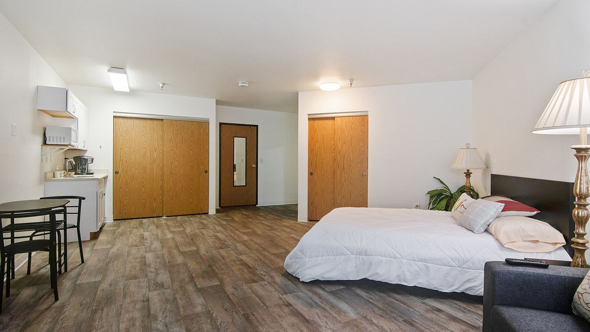 A spacious minimalist bedroom with wooden floors and white bedding, featuring closet doors and a mirror.