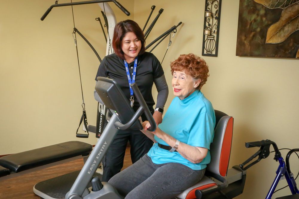 An elderly woman exercising on gym equipment with a trainer assisting her.