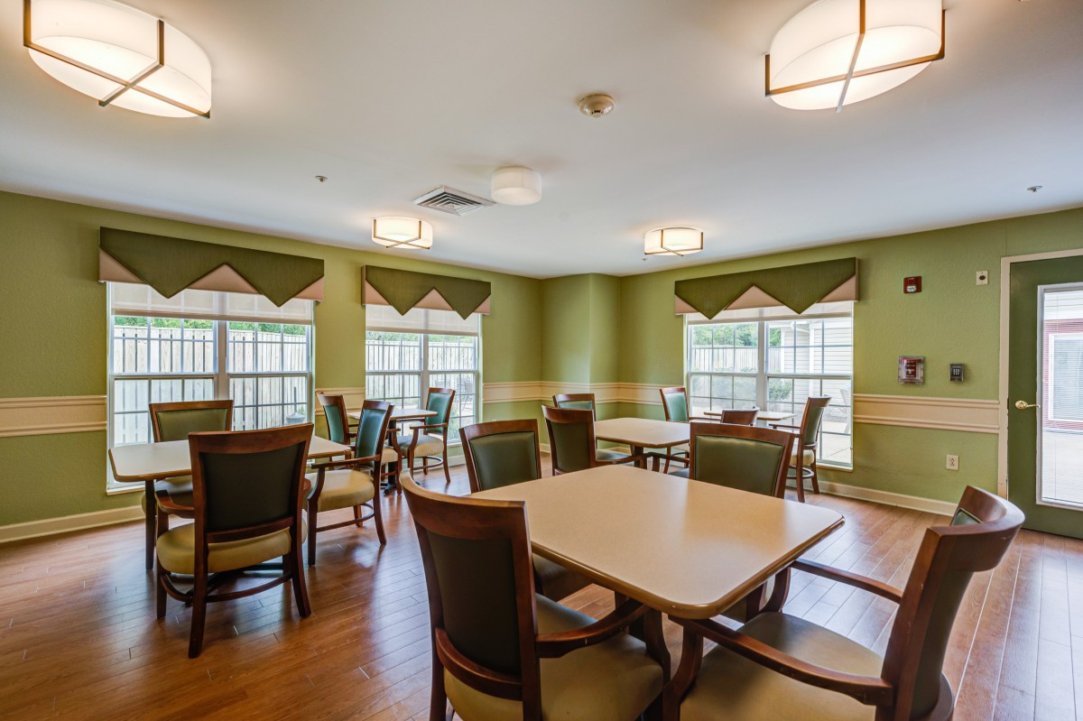A well-lit dining room with multiple tables and chairs, featuring green walls and large windows.