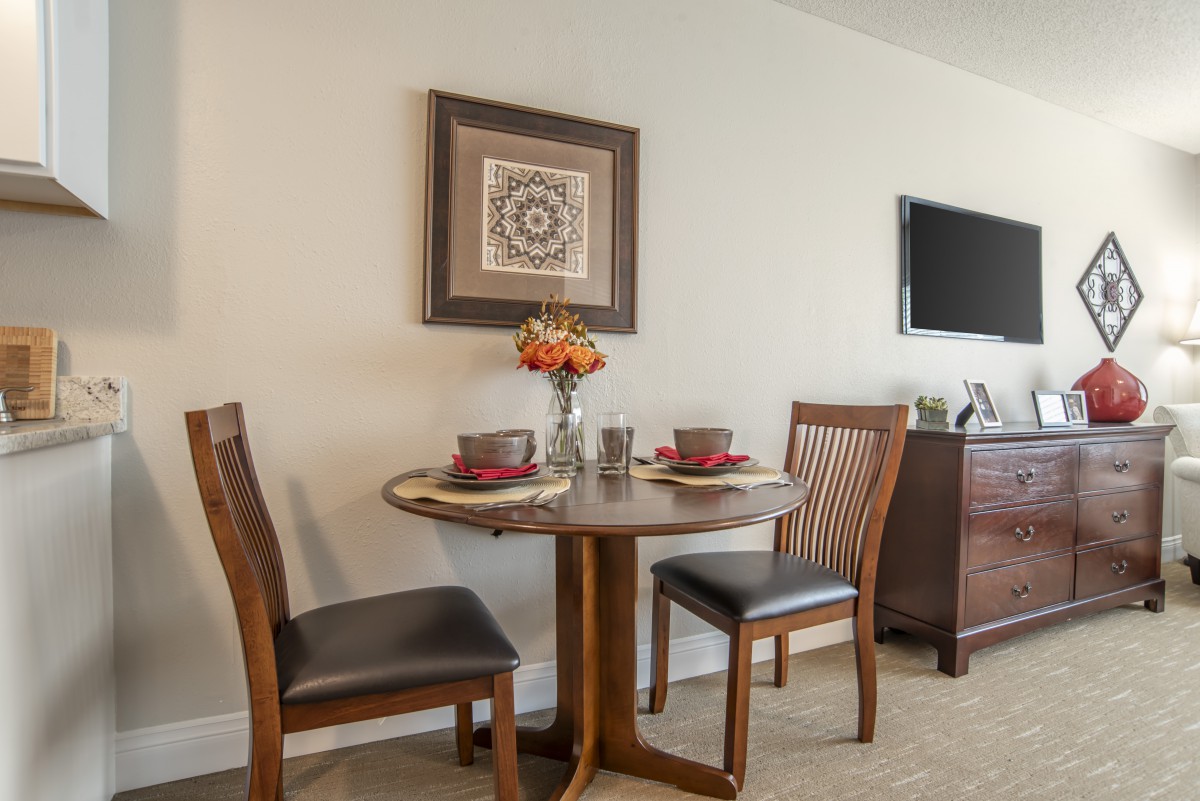 A cozy dining area with a round table set for two, featuring elegant chairs and a decorative vase of flowers, enhanced by wall art and a flat-screen TV.