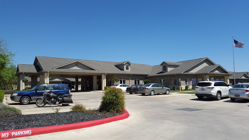 A view of a senior living facility featuring a parking area and well-maintained landscaping under a clear blue sky.