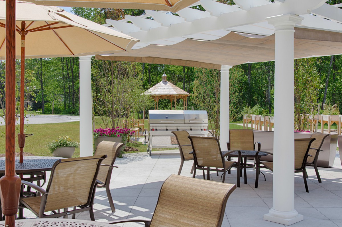 A serene outdoor patio with chairs and tables under a pergola, featuring a grill in the background.