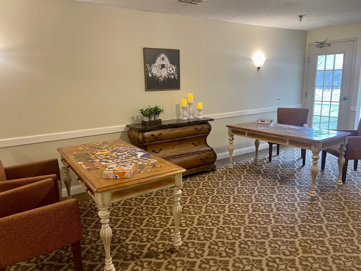 A well-furnished room with two tables, chairs, and a decorative carpet, featuring a painting on the wall and a flower vase on the chest of drawers.