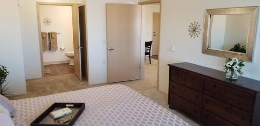 A cozy bedroom with a neatly made bed and a wooden dresser, leading to an open doorway.
