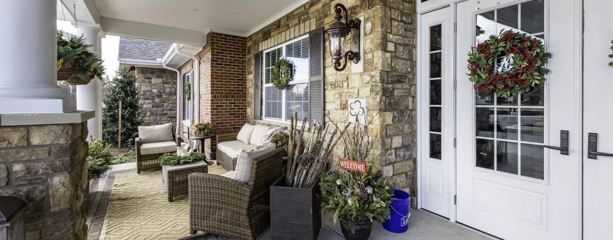 A cozy porch featuring wicker furniture, decorative plants, and a wreath on the window against a brick and stone wall.