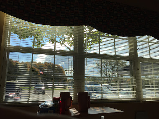 A view through a window with blinds, showcasing trees and a partly cloudy sky.