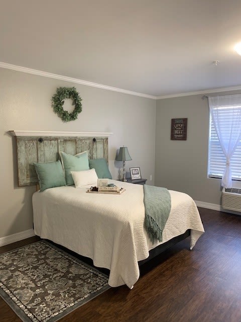 A cozy bedroom with soft green and white decor, featuring a bed with pillows, wall art, and a window letting in natural light.